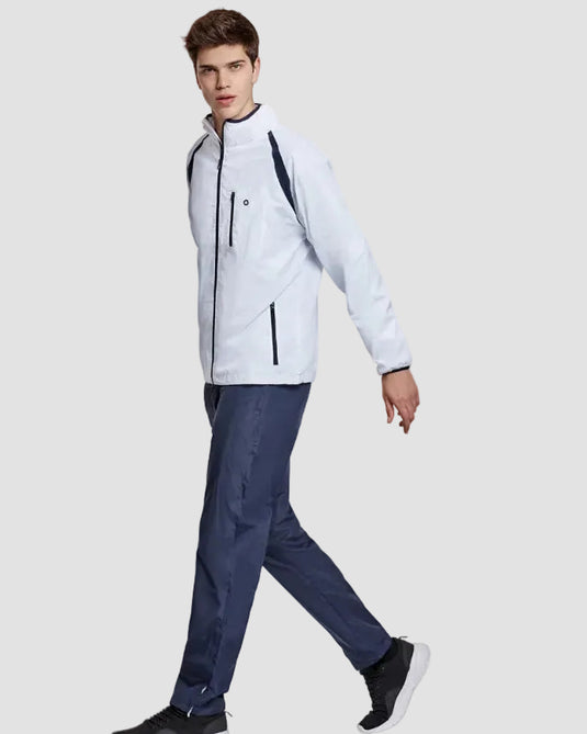 A man wearing a light gray and navy blue athletic jacket, dark blue pants, and black sneakers, walking against a plain light gray studio background.