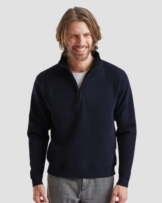 A smiling man with shoulder-length brown hair models a navy blue quarter-zip pullover sweater over a light gray t-shirt and gray jeans, set against a solid light gray background.