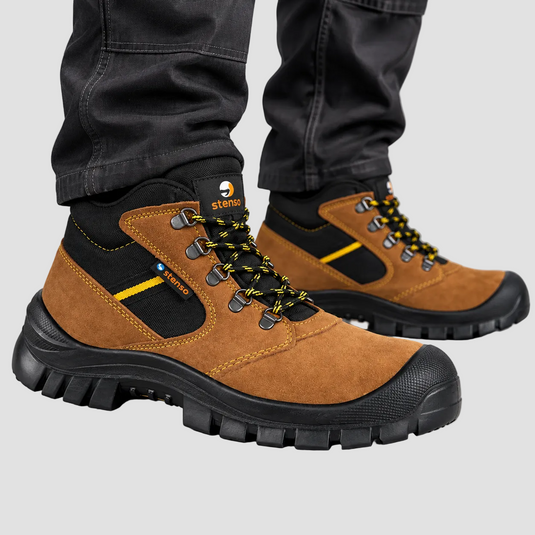 A pair of brown and black Sturdi work boots with yellow accents, shown on a person wearing dark gray pants against a light gray studio background.