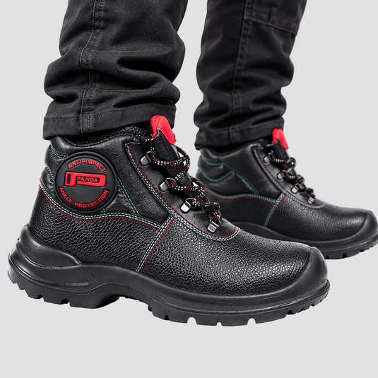 Black leather safety boots with red accents and green stitching, shown on a person wearing black jeans against a light gray studio background.