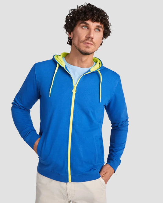 A man wearing a bright blue zip-up hoodie with neon yellow-green contrast details on the hood lining, zipper, and drawstrings, styled over a light blue t-shirt and off-white trousers, against a plain light gray studio background.