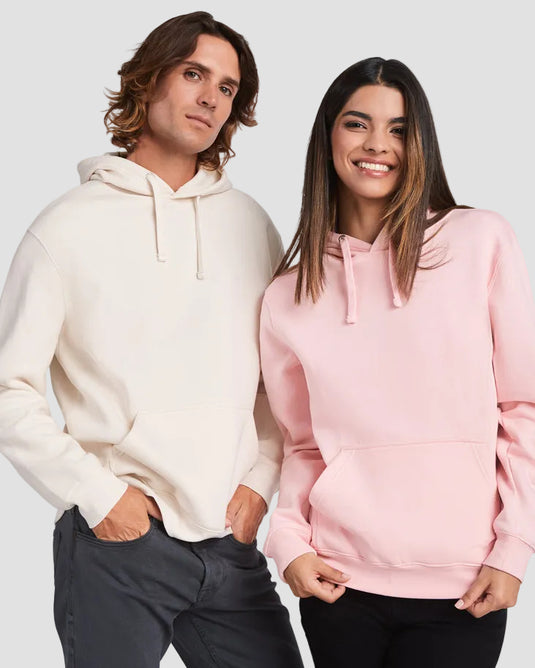 A man and a woman modeling matching pullover hoodies, with the man wearing an off-white hoodie and the woman wearing a light pink hoodie, set against a plain light gray studio background.