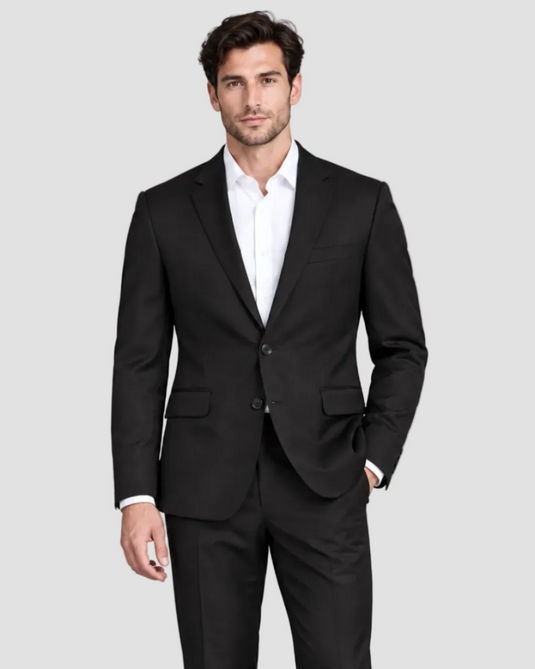 A man modeling a black two-piece suit with a white dress shirt, standing against a solid gray studio background.