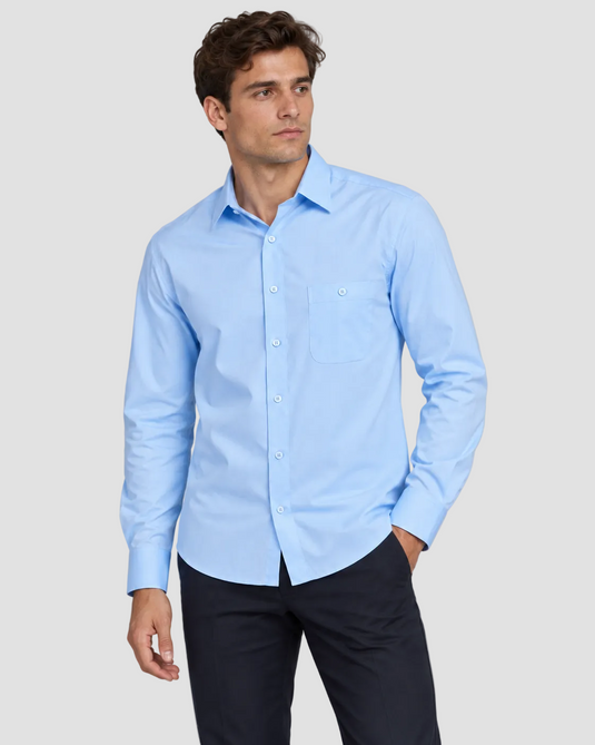 A man wearing a light blue long-sleeve button-down shirt with a single chest pocket, paired with dark trousers, standing against a solid gray studio background.
