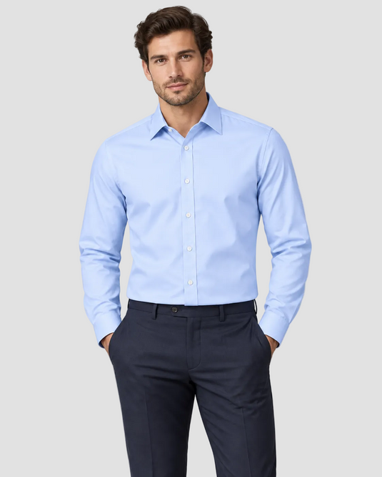 A man wearing a light blue long-sleeve dress shirt and dark navy trousers, standing against a light gray studio background.