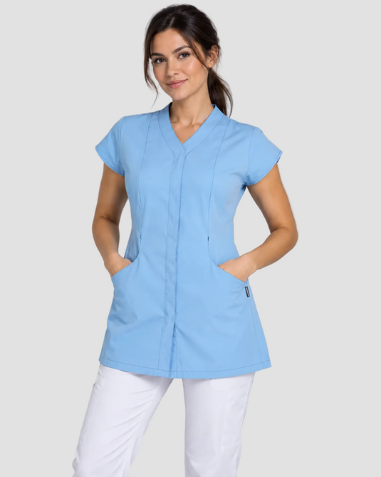 A woman modeling a light blue scrub top with short sleeves, a V-neck, and front pockets, paired with white pants, against a solid gray studio background.