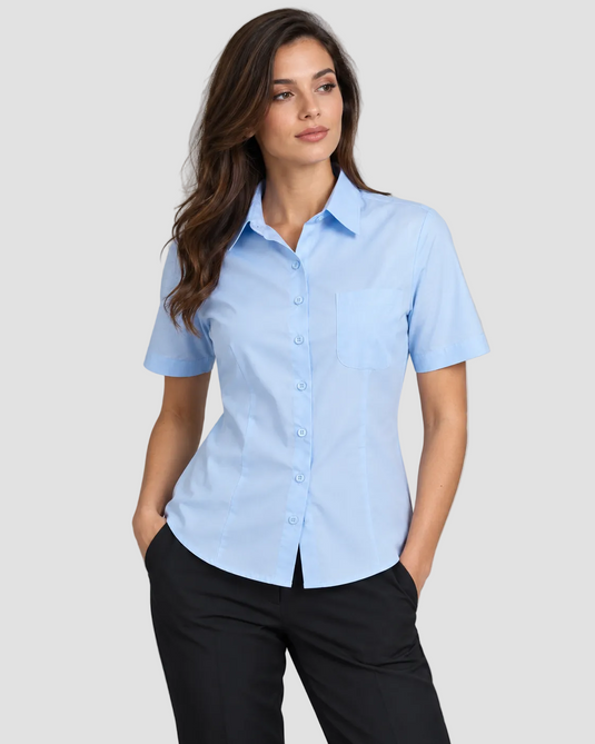 A woman modeling a light blue short-sleeve button-up shirt with a chest pocket, paired with black pants, against a solid gray studio background.
