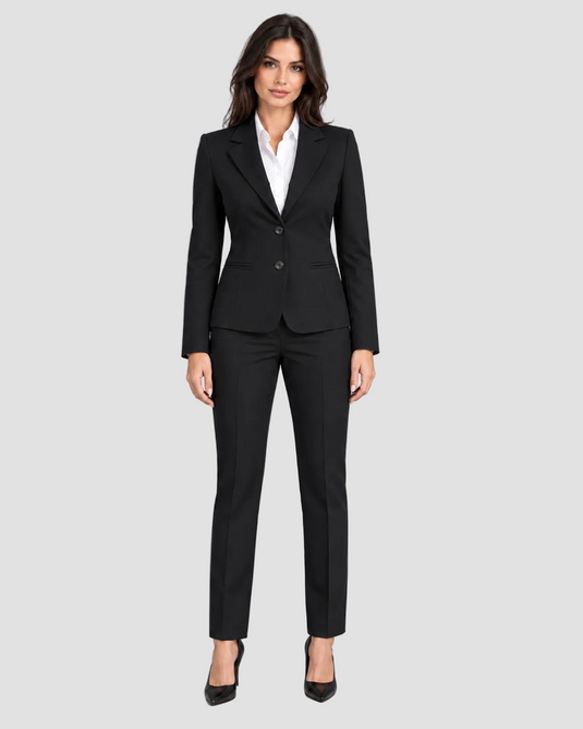 A woman modeling a black tailored two-piece suit with a white collared shirt and black pointed heels against a light gray studio background.
