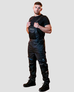 A man wearing black overalls over a black t-shirt and black boots, standing in a brick archway corridor with warm lighting.
